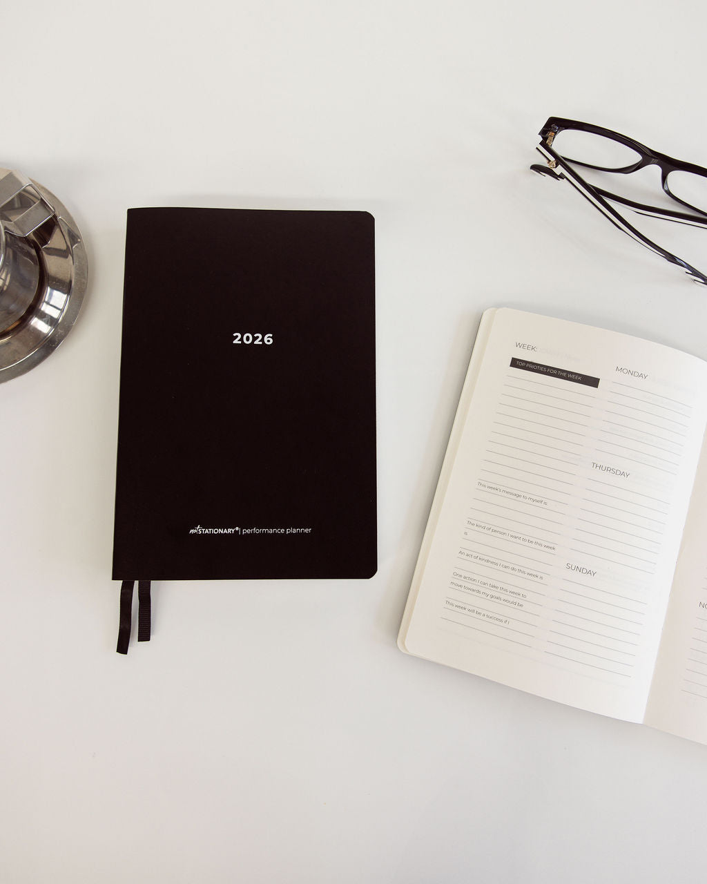 Black planner for 2026 on a white surface with an open weekly planner and glasses.