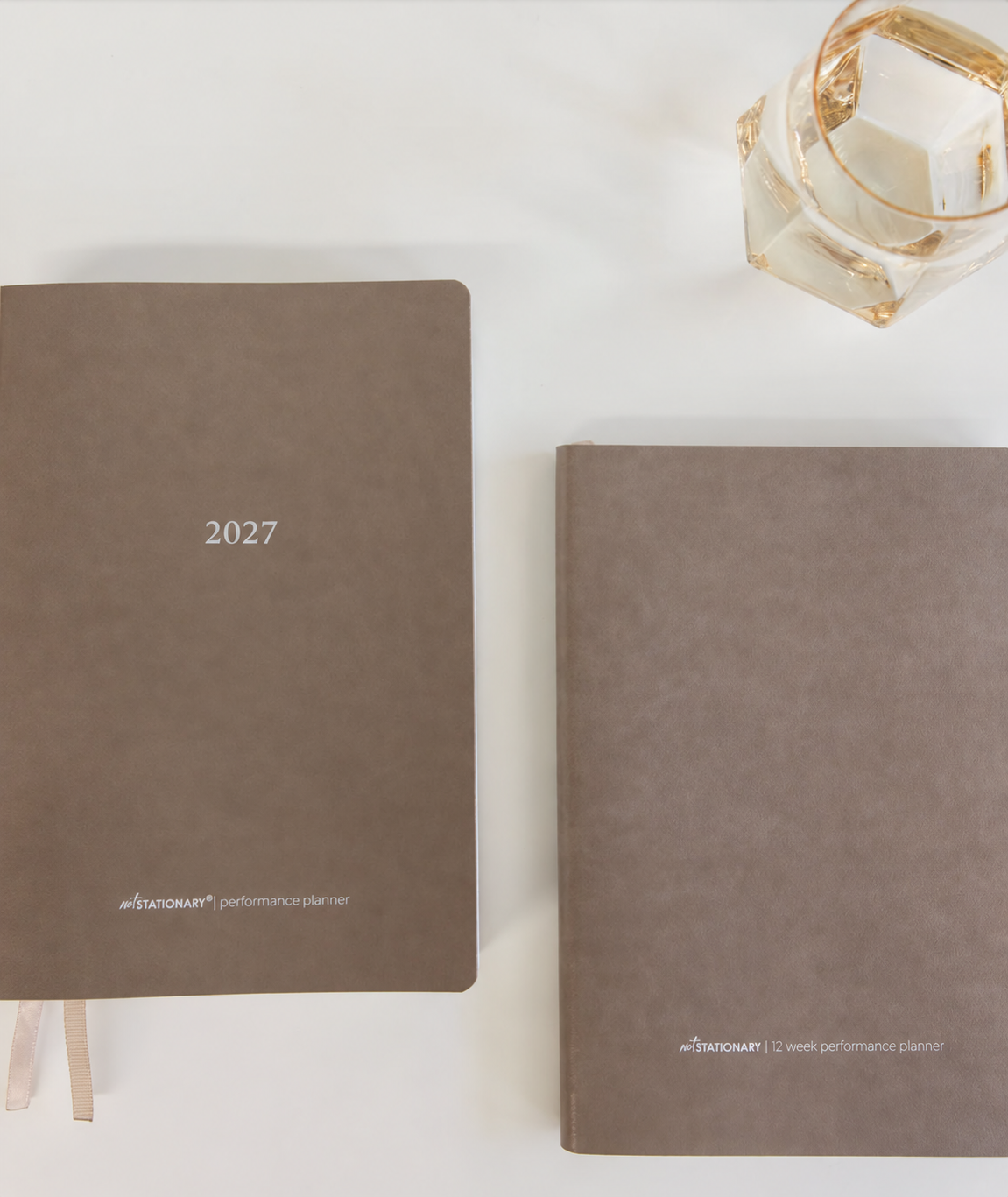 Two beige / stone vegan leather planners on a white surface, one standing upright labeled '2027' and one lying flat with the front cover visible. A faceted glass is partially visible in the top-right corner.