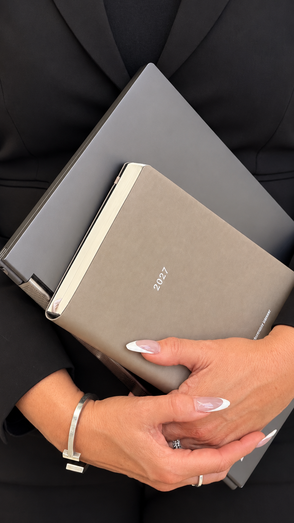 A close-up of a person in a black blazer holding a stone 2027 performance planner under their arm. The person has manicured nails with a French tip style and is wearing a silver bracelet.