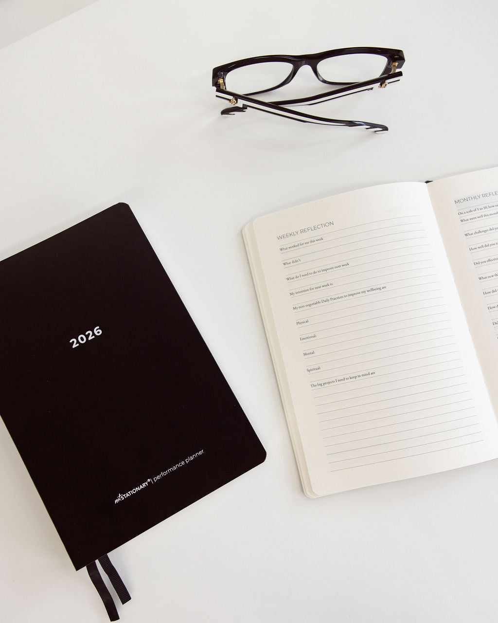Black planner with '2026' and an open notebook with reflection prompts on a white surface.