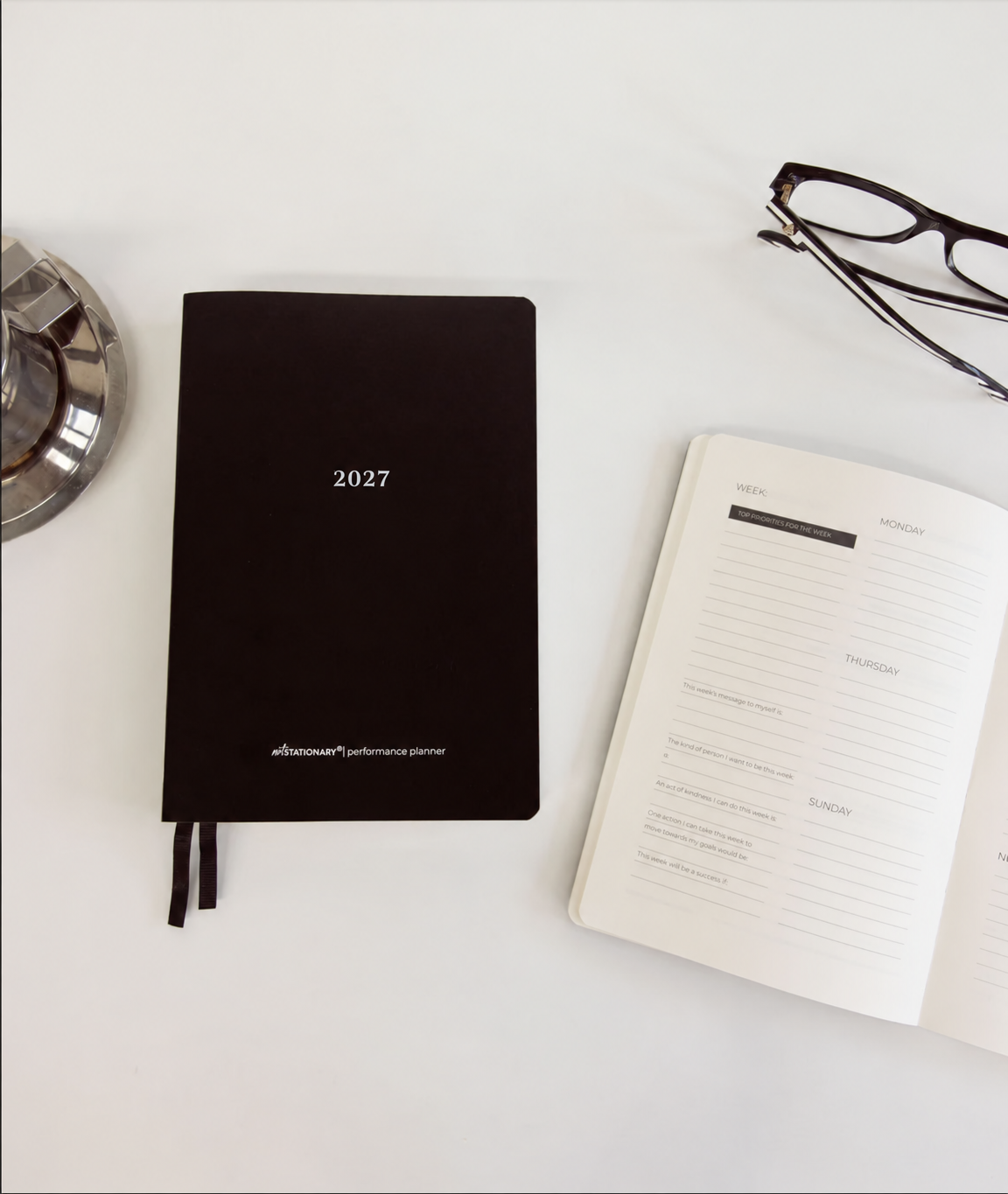 A black vegan leather 2027 planner lies open on a white surface next to an open white daily planner and a pair of black-framed glasses.