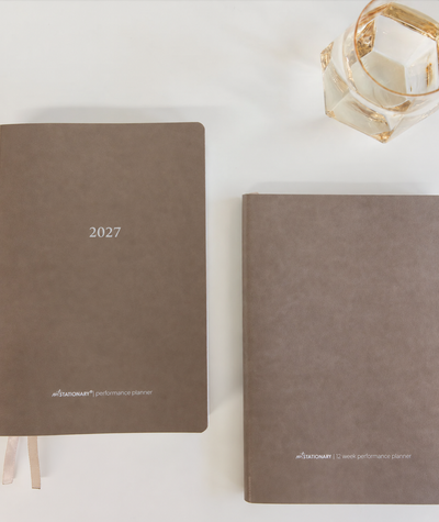 Two beige / stone vegan leather planners on a white surface, one standing upright labeled '2027' and one lying flat with the front cover visible. A faceted glass is partially visible in the top-right corner.
