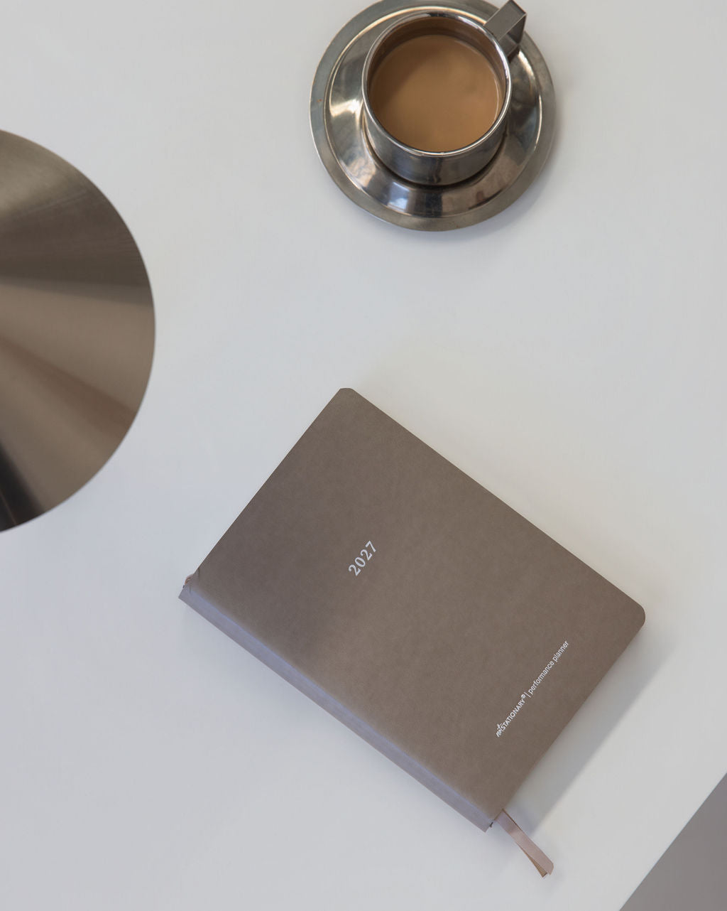 A taupe 2027 daily planner with a matching ribbon bookmark lies on a white desk next to a stainless steel coffee cup and saucer.