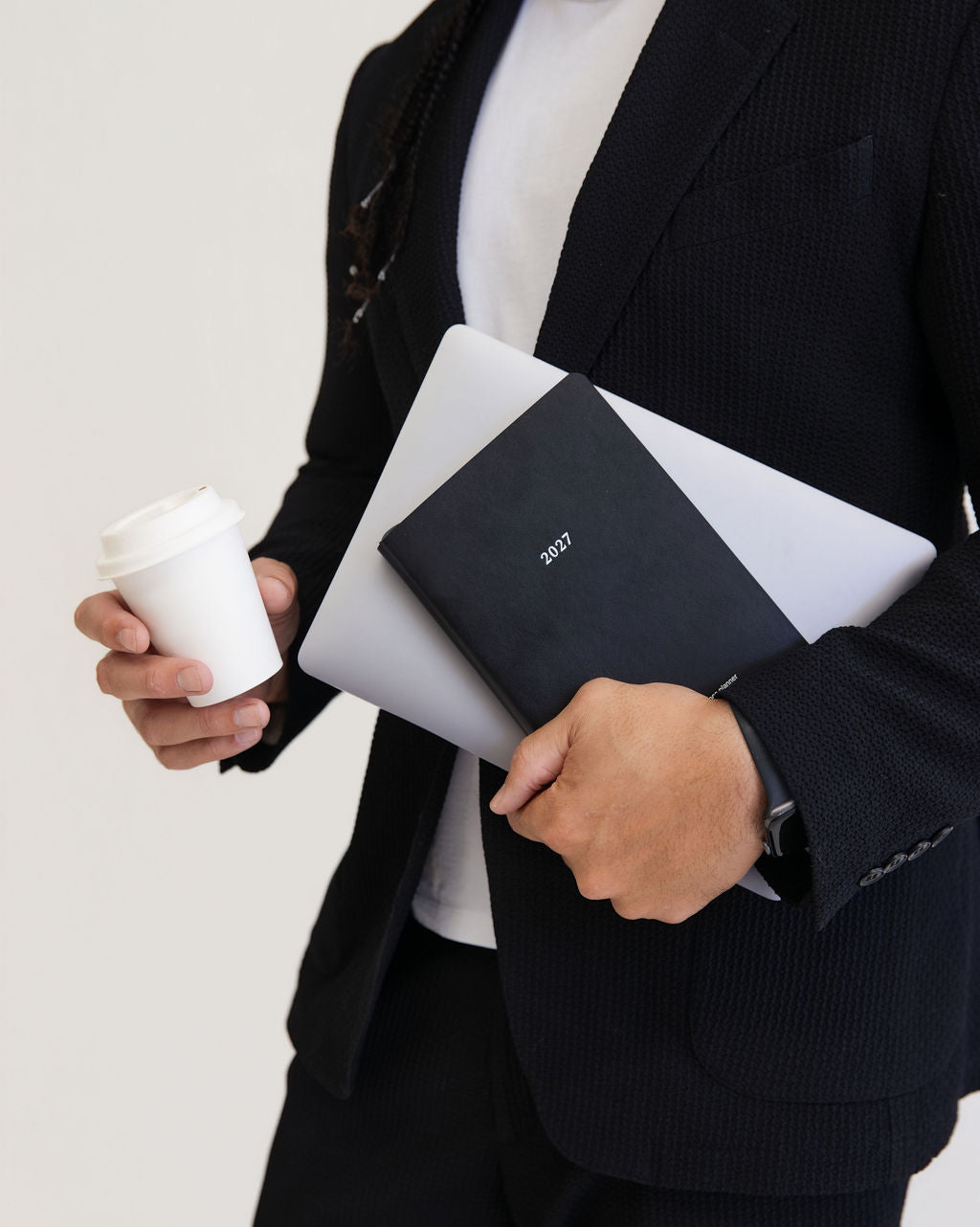 A person in a dark blazer holding a black 2023 planner, a silver laptop, and a white coffee cup against a plain light background.