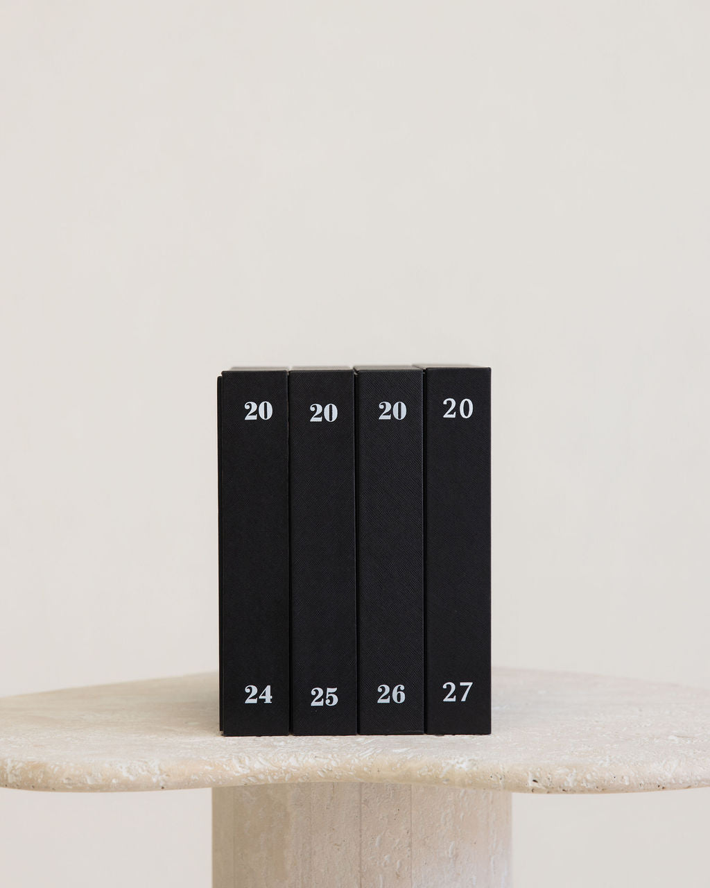 Four slim black books or boxes standing upright on a light stone surface against a warm off-white background. Each item is marked with the number 20 near the top and a sequential number from 24 to 27 near the bottom.