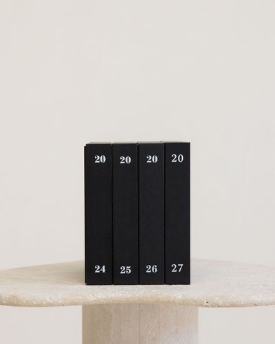 Four slim black books or boxes standing upright on a light stone surface against a warm off-white background. Each item is marked with the number 20 near the top and a sequential number from 24 to 27 near the bottom.