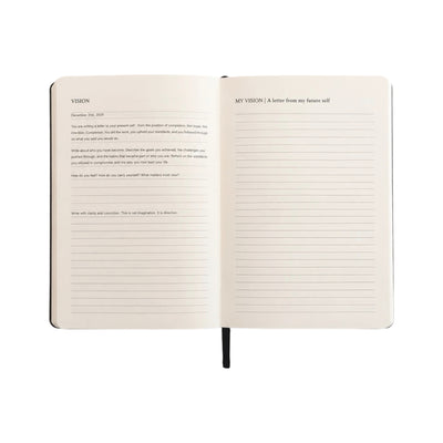 An open notebook showing two cream-colored pages with lined writing space and printed headings, with a black ribbon bookmark hanging from the bottom center on a plain white background.