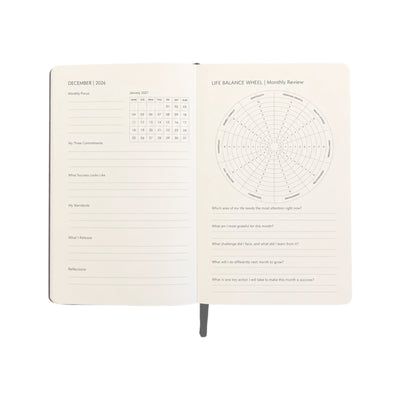 An open cream-colored planner or journal showing two interior pages: a monthly calendar and a life balance wheel with reflection prompts, photographed on a plain white background.