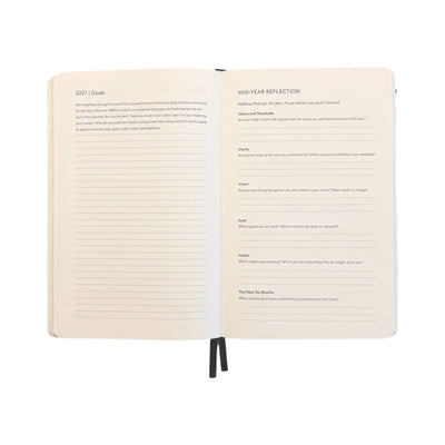 An open hardcover journal showing two interior pages with writing guides titled '2027 Goals' and 'Mid-Year Reflection,' photographed on a plain white background.